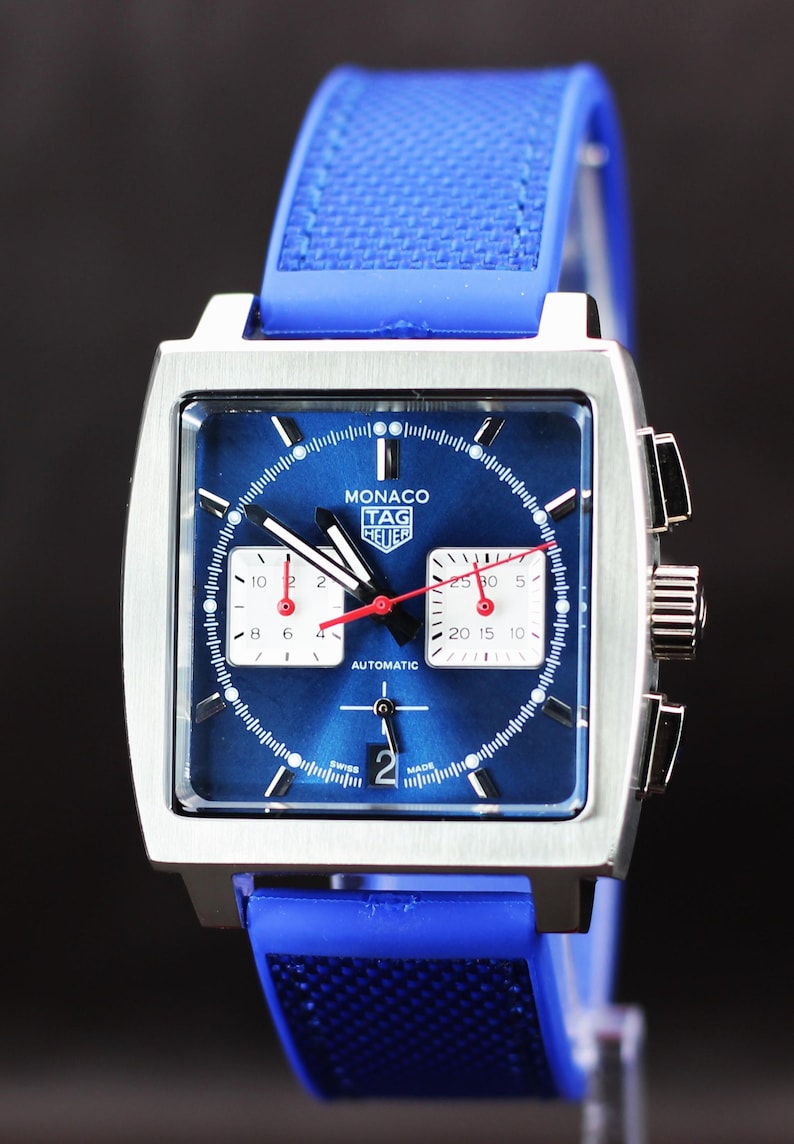 May include: A square-faced wristwatch with a blue dial and a blue rubber strap. The watch features white subdials, red hands, and the text "MONACO TAG HEUER AUTOMATIC". The watch is made in Switzerland.