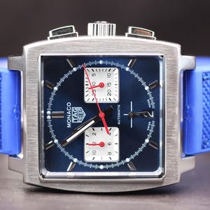 May include: A square-faced TAG Heuer Monaco watch with a blue dial and a blue rubber strap. The watch has a silver-colored case and white sub-dials with red hands. The watch face displays the word "Monaco" and the TAG Heuer logo.
