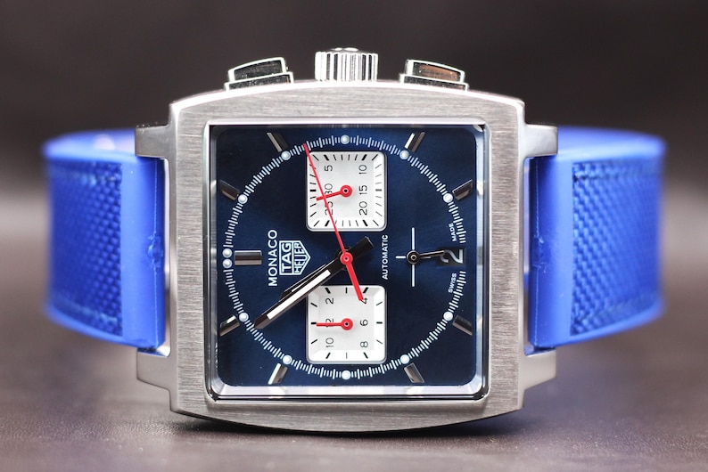 May include: A square-faced TAG Heuer Monaco watch with a blue dial and a blue rubber strap. The watch has a silver-colored case and white sub-dials with red hands. The watch face displays the word "Monaco" and the TAG Heuer logo.