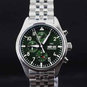 May include: A silver-tone stainless steel chronograph wristwatch with a dark green dial, featuring white Arabic numerals, three sub-dials, a day-date window at the three o'clock position, and the text IWC SCHAFFHAUSEN printed on the watch face.