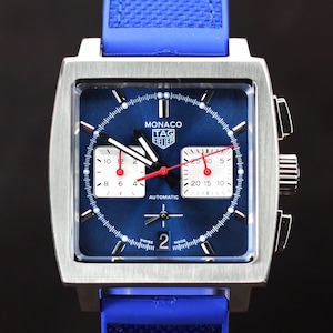 May include: A square-faced, stainless steel watch with a blue dial and blue rubber strap. The watch face features white sub-dials, red hands, and the text "MONACO TAG HEUER AUTOMATIC". The watch has a date display at the bottom.