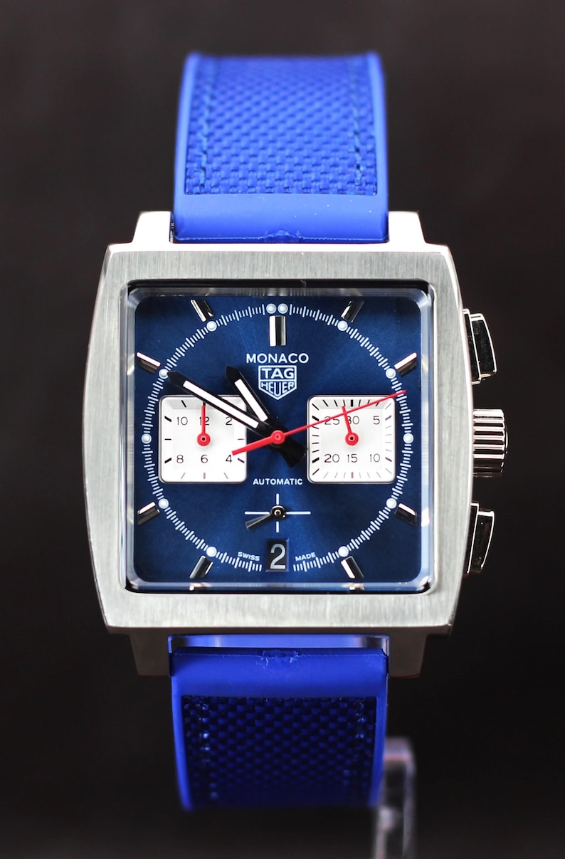 May include: A square-faced, stainless steel watch with a blue dial and blue rubber strap. The watch face features white sub-dials, red hands, and the text "MONACO TAG HEUER AUTOMATIC". The watch has a date display at the bottom.
