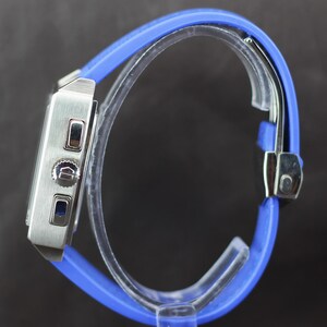 May include: A silver-toned wristwatch with a rectangular face and a vibrant blue silicone band. The watch features a stopwatch function and a secure clasp. The band is approximately 9 inches (23 cm) in length, offering a comfortable fit.