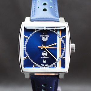 Könnte beinhalten: Eine quadratische Uhr mit blauem Zifferblatt und blauem Lederarmband. Auf dem Zifferblatt steht "MONACO TAG HEUER" und "GULF". Das Armband hat ein perforiertes Design. Das Gehäuse der Uhr ist silberfarben.