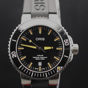Pode incluir: Um relógio de pulso Oris Aquis com mostrador preto, marcadores de hora amarelos e ponteiros amarelos. O relógio possui uma luneta giratória preta com números brancos, uma janela de data às 6 horas e uma pulseira de borracha preta com o logo Oris.