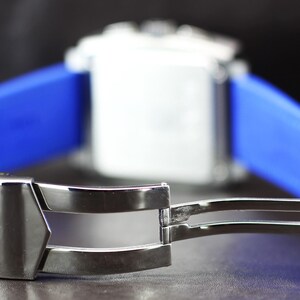 May include: Close-up of a silver watch clasp with a blue strap. The clasp is open, revealing its intricate design. The watch face is white, and the strap is a vibrant blue. The background is dark, highlighting the watch's details.