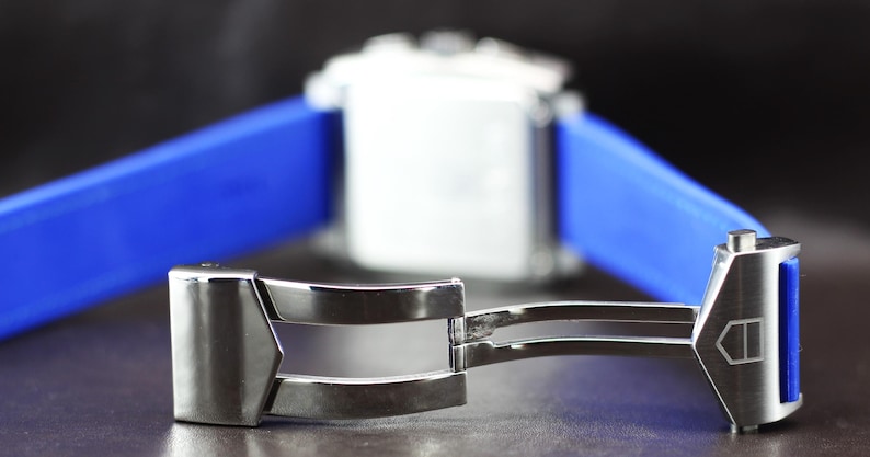 May include: Close-up of a silver watch clasp with a blue strap. The clasp is open, revealing its intricate design. The watch face is white, and the strap is a vibrant blue. The background is dark, highlighting the watch's details.