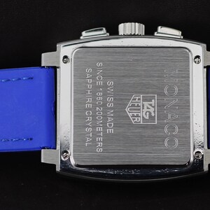 May include: A silver-toned Monaco watch with a square face and blue leather strap. The watch back features the text "SWISS MADE" and "SAPPHIRE CRYSTAL". The blue strap has the brand name "TAG Heuer" embossed on it.