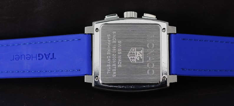 May include: A silver-toned Monaco watch with a square face and blue leather strap. The watch back features the text "SWISS MADE" and "SAPPHIRE CRYSTAL". The blue strap has the brand name "TAG Heuer" embossed on it.