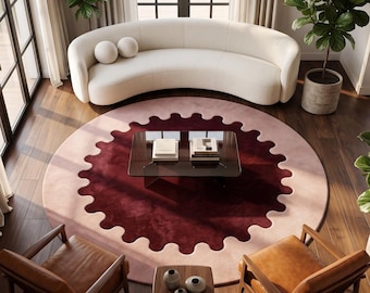 Op de afbeelding: Een groot rond vloerkleed met een diepbordeauxrood middenstuk met een geschulpte rand, omringd door een brede rand van zacht lichtroze. Het kleed ligt op een donkere houten vloer onder een glazen salontafel met twee boeken erop.