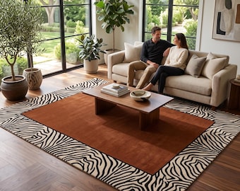 Op de afbeelding: Een rechthoekig vloerkleed met een effen, gebrand oranje middenpaneel, omlijst door een brede rand met een zwart-wit zebrastreeppatroon. Het kleed ligt op een donkere houten vloer in een kamer met grote ramen en een lichtgekleurde bank.