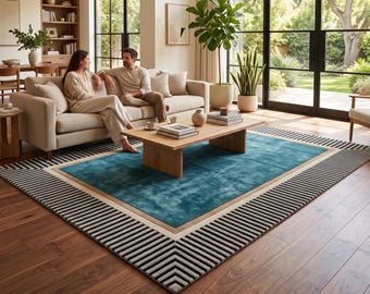 Op de afbeelding: Een rechthoekig vloerkleed met een effen blauwgroen middenpaneel, een beige rand en een buitenframe met een zwart-wit gestreept chevronpatroon. Het kleed ligt op een donkere houten vloer onder een houten salontafel in een woonkamer.