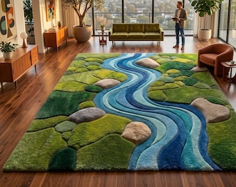 May include: A rectangular area rug featuring a textured, three-dimensional landscape design. The rug displays a winding blue river flowing through patches of green mossy terrain, accented by scattered smooth, light-colored stone shapes.
