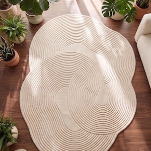 May include: A large, cream-colored area rug with a unique, abstract design. The rug features a series of concentric, curved lines, creating a textured, wave-like pattern. The rug is placed on a dark wood floor, surrounded by potted plants.