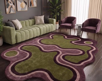 Op de afbeelding: Een vloerkleed met een onregelmatige, organische vorm met gelaagde, golvende banen in olijfgroen, donkerpruim en zacht mauve. Het kleed heeft een pluche, getextureerd oppervlak en ligt op een donkere houten vloer met een veelkleurig patroon.