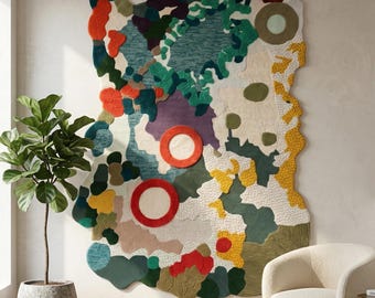 Op de afbeelding: Een groot, abstract wandkleed met een kleurrijk, organisch ontwerp. Het kleed heeft een mix van texturen en kleuren, waaronder teal, groen, rood en geel. Twee grote rode cirkels zijn prominent aanwezig. Een plant in een pot en een crèmekleurige stoel staan op de voorgrond.