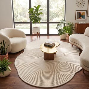 Könnte beinhalten: Ein cremefarbener, abstrakt geformter Teppich mit einem strukturierten, geriffelten Design dominiert das Wohnzimmer. Ein goldfarbener Couchtisch steht auf dem Teppich, und ein geschwungenes Sofa und ein Sessel befinden sich in der Nähe. Der Raum ist voller Pflanzen und natürlichem Licht.