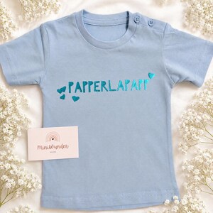 Könnte beinhalten: Ein hellblaues Kurzarm-T-Shirt mit dem Schriftzug "PAPPERLAPAPP" in metallisch türkiser Schrift, verziert mit kleinen türkisen Herzen. Das Shirt hat zwei Knöpfe am Schulterausschnitt und eine kleine rechteckige Karte mit der Aufschrift "Miniwunder".