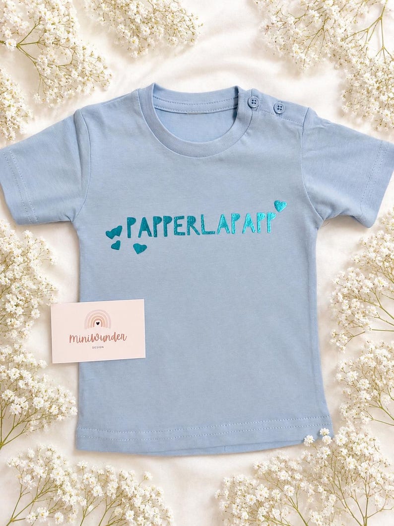 May include: A light blue short-sleeved t-shirt featuring the text "PAPPERLAPAPP" in a metallic teal font, accented with small teal hearts. The shirt includes two buttons on the shoulder neckline and a small rectangular card labelled "Miniwunder".