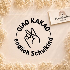May include: A rectangular white card featuring a black circular graphic with the text "CIAO KAKAO endlich Schulkind" and a hand gesture showing two fingers. A smaller rectangular tag with the "MiniWunder DESIGN" logo and a rainbow icon is placed above the main card.