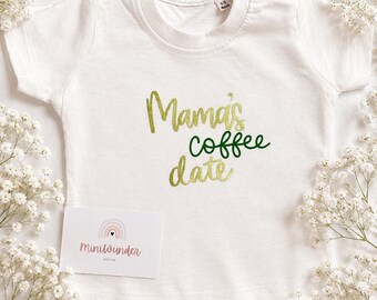May include: A white short-sleeved baby t-shirt with the text Mama's coffee date printed in metallic gold and dark green. The shirt features a small tag at the neckline and is accompanied by a rectangular white card with the text Miniwunder design.