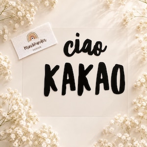 May include: A clear rectangular acrylic sign featuring the black text ciao KAKAO in a bold, rounded font. A small white fabric tag with a rainbow graphic and the text MiniWunder DESIGN is positioned in the upper left corner of the sign.