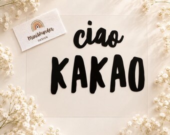 May include: A clear rectangular acrylic sign featuring the black text ciao KAKAO in a bold, rounded font. A small white fabric tag with a rainbow graphic and the text MiniWunder DESIGN is positioned in the upper left corner of the sign.