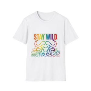 Op de afbeelding: Wit T-shirt met een regenboogkleurige afbeelding van een buffelkop en de tekst "STAY WILD". Het ontwerp bevat een landschap met bomen, planten en een wereldbol. Ronde hals.