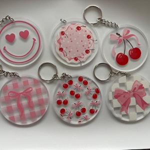 May include: A collection of six clear acrylic keychains with silver key rings. Each keychain features a different pink design: a smiley face with hearts, cherries, bows, and a gingham pattern. The keychains are round.