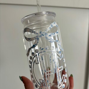 Pode incluir: Copo de vidro transparente com tampa e canudo. O copo apresenta um laço decorativo e um design floral em tons de azul e branco. O design inclui uma letra "H" estilizada.