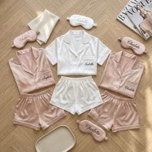 May include: Three sets of satin pyjamas in white and dusty pink, each featuring a button-down shirt with a chest pocket and matching shorts. The garments are embroidered with the names Sophia, Isabella, and Charlotte, accompanied by matching eye masks.