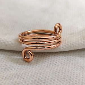 Puede incluir: Un anillo de metal color cobre con un diseño de múltiples espirales. La banda consta de tres bucles continuos de alambre que se envuelven para formar una espiral, con cada extremo del alambre terminando en un pequeño bucle circular.