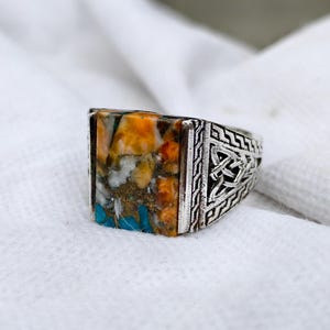 Puede incluir: Un anillo de plata con una incrustación rectangular de piedra multicolor, que incluye segmentos de color naranja, blanco y turquesa. La banda de plata está decorada con intrincados patrones geométricos en relieve y un diseño de nudo.