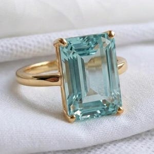 May include: A gold ring featuring a large, rectangular, emerald-cut light blue gemstone held in place by four prongs. The band is smooth and polished, showcasing a simple design that highlights the clarity and pale blue color of the stone.