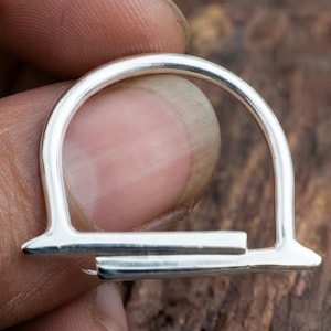 Peut inclure: Une bague de couleur argentée présentant un arc supérieur semi-circulaire relié à une base droite et horizontale. Le métal possède une finition lisse et polie avec une forme géométrique distincte créant un espace ouvert dans la structure.