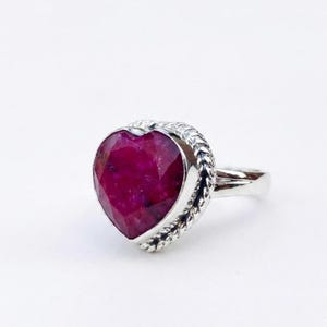 Può includere: Un anello in argento con una gemma sfaccettata a forma di cuore di colore rosa intenso, incastonata in un bordo metallico a corda intrecciata. Il gambo è liscio e lucido, collegato alla montatura a cuore che mostra inclusioni naturali.