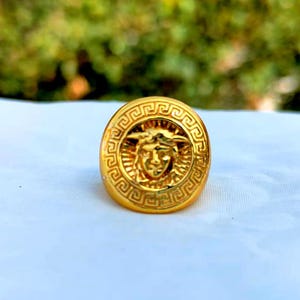 May include: A gold-toned ring featuring a circular face with a raised relief of a face at the center, surrounded by a detailed Greek key pattern border. The metallic surface has a polished finish that reflects light across the circular design.
