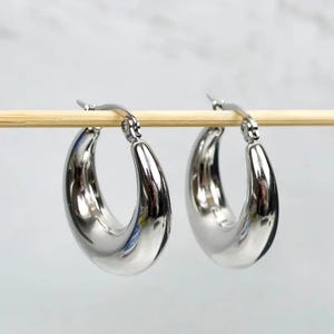 Può includere: Un paio di orecchini a cerchio in tonalità argento lucido con un design spesso, arrotondato e a forma di mezzaluna. Ogni orecchino presenta un meccanismo di chiusura a cerniera sulla parte superiore, creando una superficie liscia.
