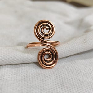 May include: A copper ring featuring a double spiral design. The metal wire is coiled into two circular shapes, one positioned above the other, connected by a central band that forms the ring structure. The finish is a polished metallic copper tone.