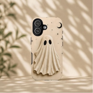 Puede incluir: Una funda de teléfono de color beige con una figura de fantasma en relieve tridimensional en el centro. El fondo de la funda está decorado con pequeñas estrellas negras y una luna creciente. La abertura de la cámara está arriba a la izquierda.