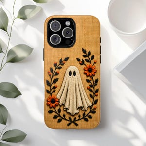 Puede incluir: Una funda de teléfono color amarillo mostaza con una superficie texturizada similar a la tela. El diseño presenta un fantasma color crema en el centro, rodeado por una corona de enredaderas negras, bayas naranjas y flores brillantes.