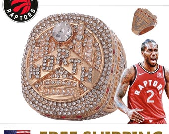 Op de afbeelding: Een goudkleurige kampioensring met het woord "NORTH" en een grote heldere steen. De ring is ingelegd met talrijke kleine, heldere stenen. Het logo van de Toronto Raptors is zichtbaar.