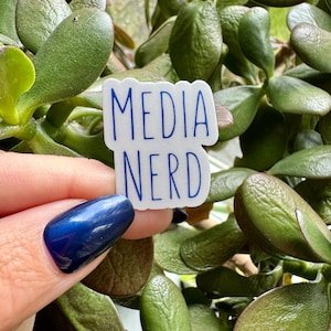 May include: A white sticker with the words "MEDIA NERD" in blue, held against a backdrop of green leaves. The sticker has a blue border and is slightly angled. The image is well-lit and in focus.