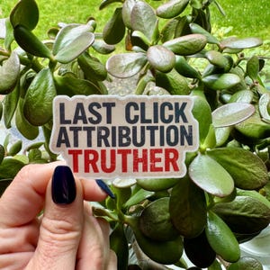 May include: A sticker with the text "LAST CLICK ATTRIBUTION TRUTHER" in black and red. The sticker is held in front of a green plant with oval-shaped leaves. The background is blurred.