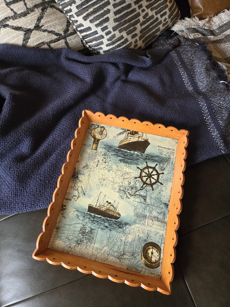 May include: Wooden serving tray with a nautical theme. The tray features a map design with images of ships, a compass, and a ship's wheel. The tray has a scalloped edge and is made of light-colored wood.