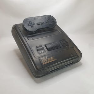 May include: A translucent black Super Nintendo Entertainment System console with a matching translucent black game controller resting on top. The console features the Nintendo Super Nintendo Entertainment System logo in gold text on the front panel.