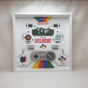May include: A white shadow box frame containing a disassembled Super Nintendo Entertainment System controller. The display features a logic board, rubber pads, triggers, a D-pad, buttons, and a controller shell, all labelled with text on a white background.