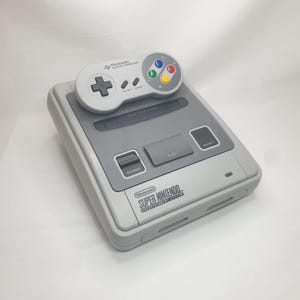 May include: A grey Super Nintendo Entertainment System console with a matching controller. The controller has a directional pad, start and select buttons, and coloured action buttons. The console has the words "Super Nintendo Entertainment System" printed on the front.