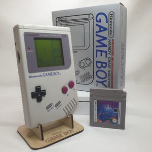 May include: A light grey Nintendo Game Boy handheld console with a green screen, black directional pad, two purple buttons, and grey Select and Start buttons. It includes a Tetris game cartridge and the original white cardboard packaging box.