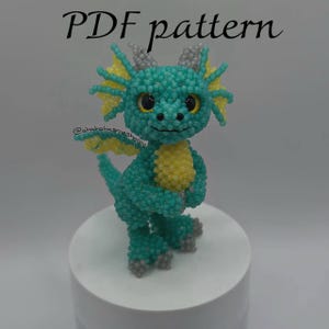 Könnte beinhalten: Eine Perlen-Drachenfigur in Türkis, Gelb und Grau. Der Drache hat große, ausdrucksstarke Augen und steht auf einem weißen Sockel. Der Text "PDF pattern" befindet sich oben im Bild.